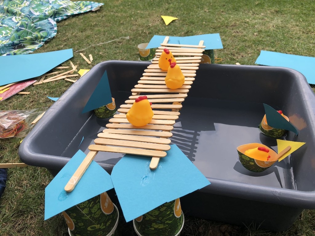 Yellow rubber chickens cross a craft-stick bridge and ride paper-cup boats in a tub of water.